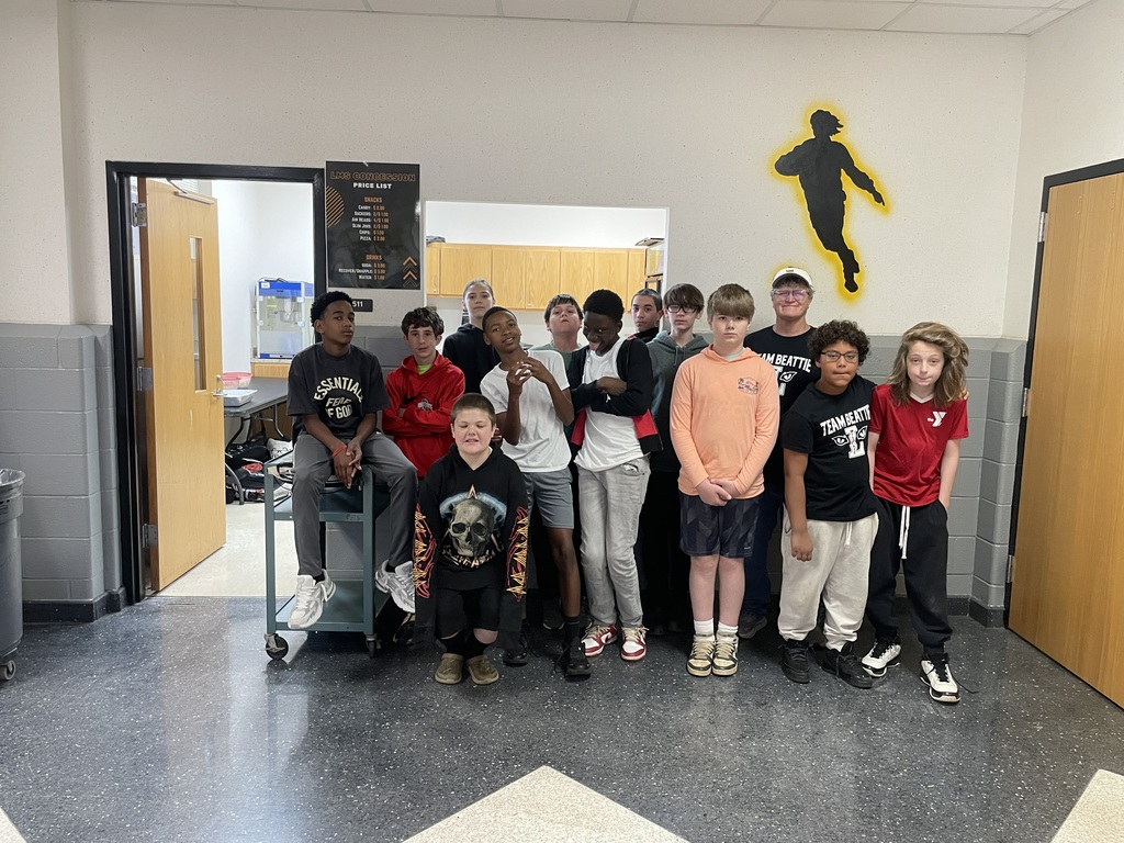 A group of twelve students and one adult posing together in a school hallway. They are standing in front of a doorway labeled 511. A yellow silhouette of a person in motion is painted on the wall behind them.