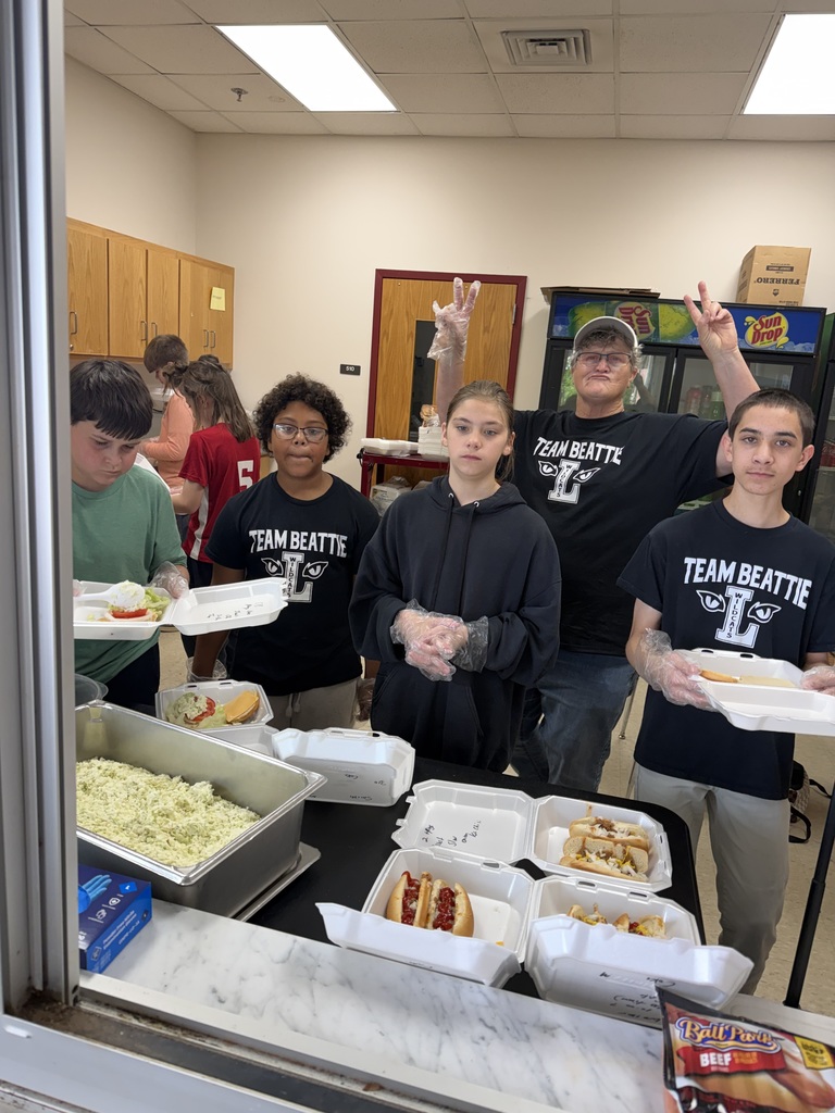 A group of four students and one adult standing behind a service counter. The students are wearing black "Team Beattie" t-shirts and clear plastic gloves. On the counter are several open foam containers with hot dogs and a large metal tray of coleslaw.