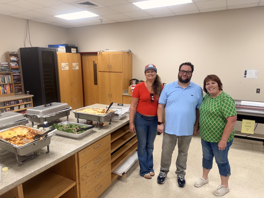 Three adults standing in a school workroom or cafeteria area behind a counter. On the counter are several silver chafing dishes containing food, including lasagna and a green salad. The room has wooden cabinets and a laminator in the background.