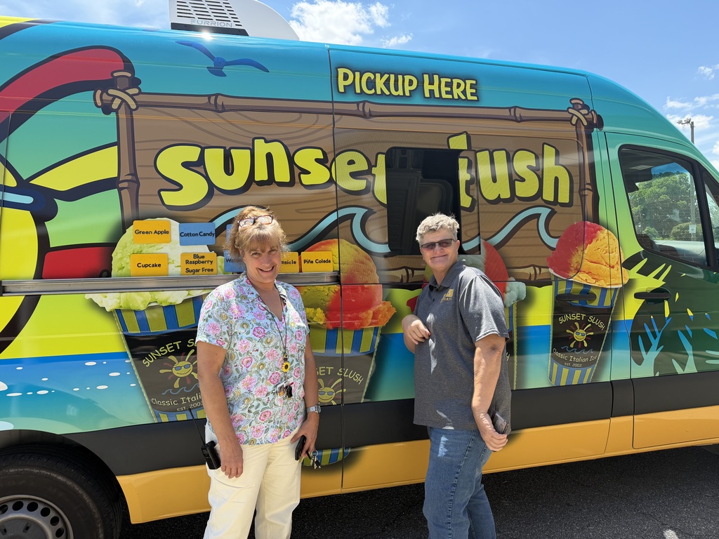Two women standing in front of a bright blue Sunset Slush food truck. The truck features a large "Sunset Slush" logo and a menu board with flavors including Green Apple, Cotton Candy, Cupcake, Blue Raspberry Sugar Free, Orange Creamsicle, and Pina Colada. A small sliding window labeled "PICKUP HERE" is visible.