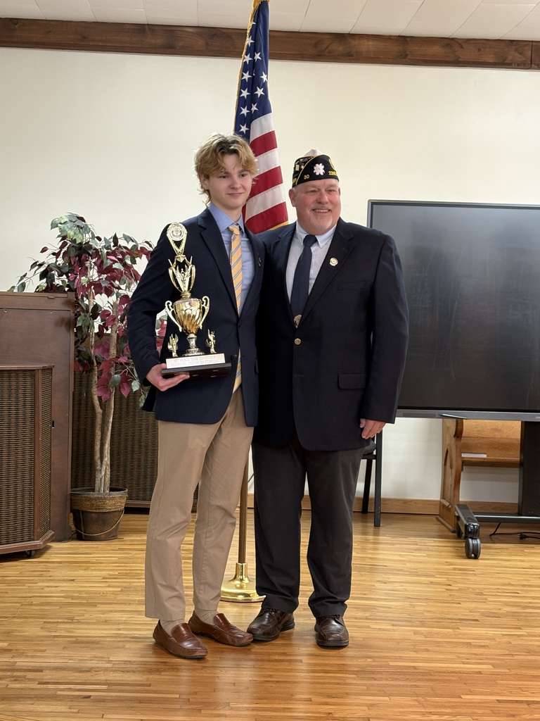 A man stands next to a student holding a trophy. A flag is behind them and a large screen to the right.