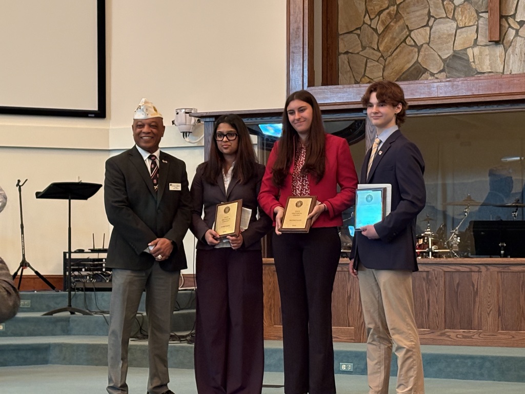 Four people stand in a church; one wears a crown. They hold certificates in their hands. Behind them, a wooden desk is on the stage.