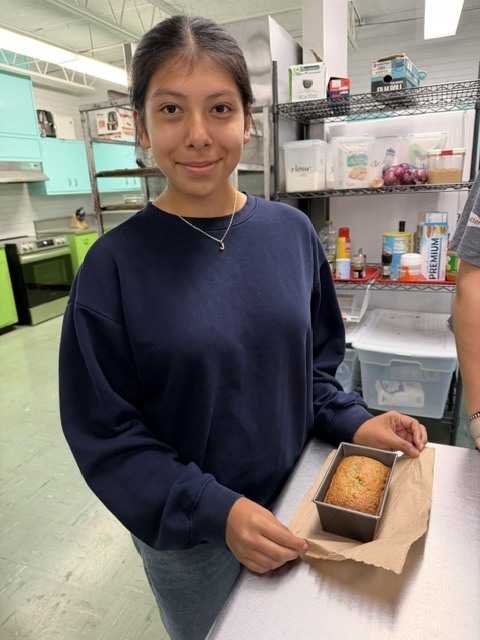 A woman in a blue shirt and jeans holds a loaf of bread wrapped in brown paper in a kitchen.