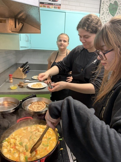 Three women in a kitchen, two using spatulas to stir food in a pan, and one holding a spoon.
