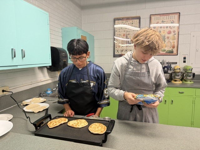 Two kids in aprons, one holding a packet, prepare pancakes on a griddle in a kitchen with blue cabinets.