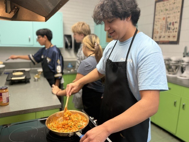 A person in a blue shirt and apron stirs food in a pan on a stove.