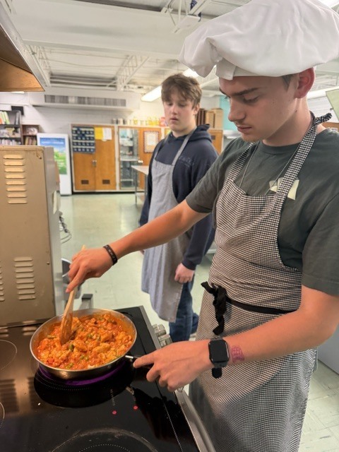 A student in a chef's hat and apron stirs a pot of food on a stove. Another student stands behind them.