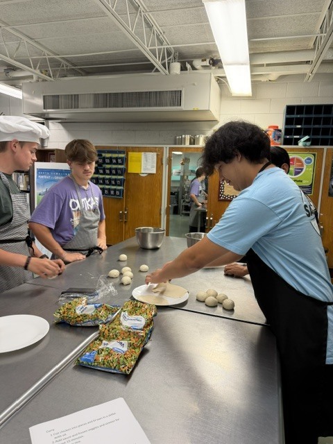 Three people wearing aprons prepare food in a kitchen. One person works with dough on a cutting board.