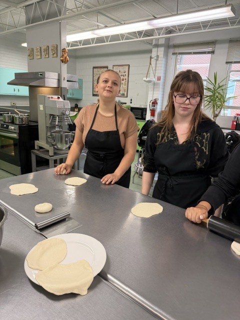 Two women in aprons prepare food in a kitchen with a table and a rolling pin.