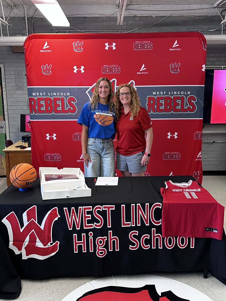 Two women stand behind a table at a sports event, one wearing a blue shirt and the other in red.