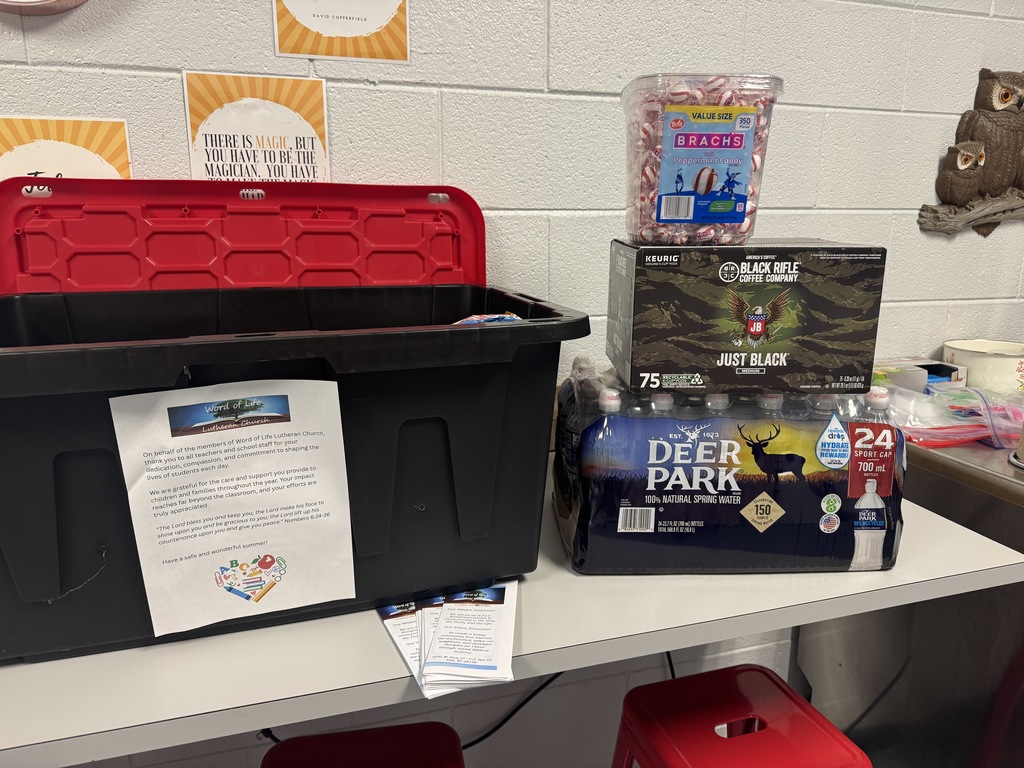 Table with two black boxes and a red one, a camouflage box, and a box of water. A bulletin board is in the background.