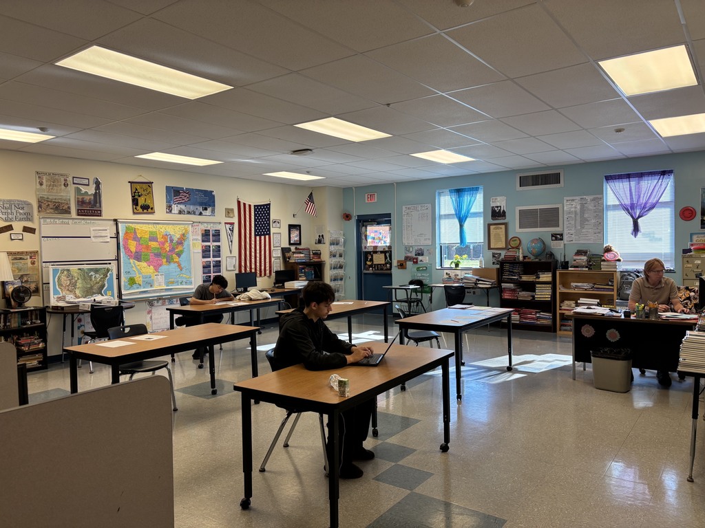 A classroom with rows of desks and chairs, some occupied. A person sits at a desk with a laptop. A map and flag are on the wall.