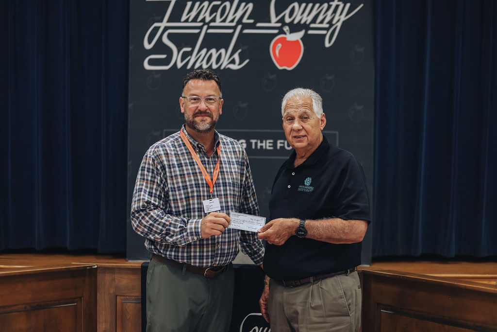 Two men stand in front of a Lincoln County Schools banner. One holds a check, the other extends their hand.