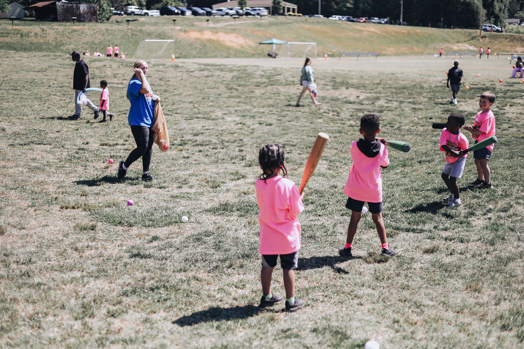 People in a grassy field play with bats and balls. A child holds a bat and looks back.
