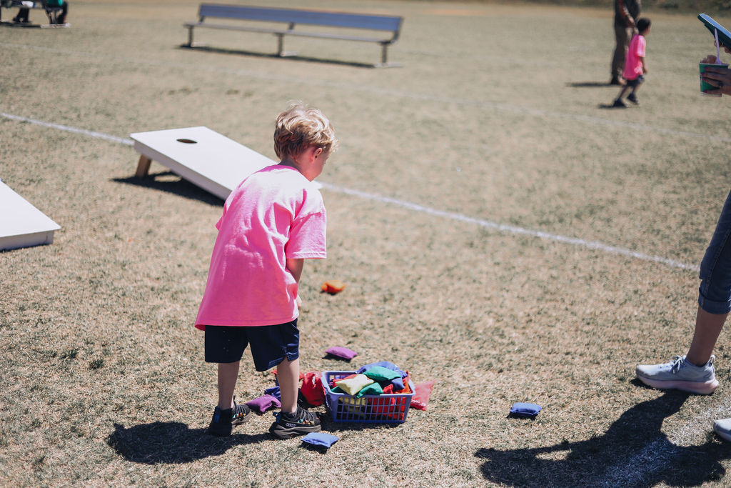 A child in a pink shirt plays with small objects on a dirt field. A white bench and cornhole boards are in the background.