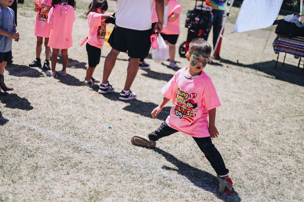 Children in pink shirts and face paint stand in a line, one kicking a ball in a sandy area.