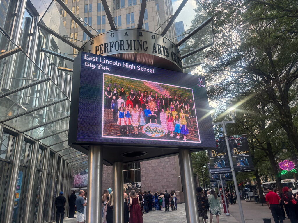 A large digital display shows a photo of students at East Lincoln High School. It stands in front of a building with many glass walls.