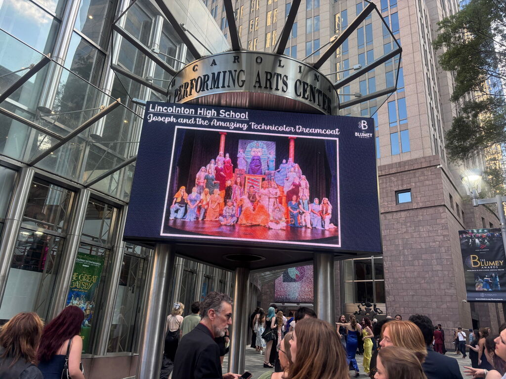 Billboard at a city center, featuring an outdoor performance by Lincoln High School's cast. The crowd stands outside a modern building.