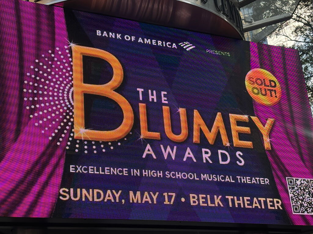 A large, purple sign with yellow text promoting "The Blumey Awards" highlights excellence in high school musical theater.