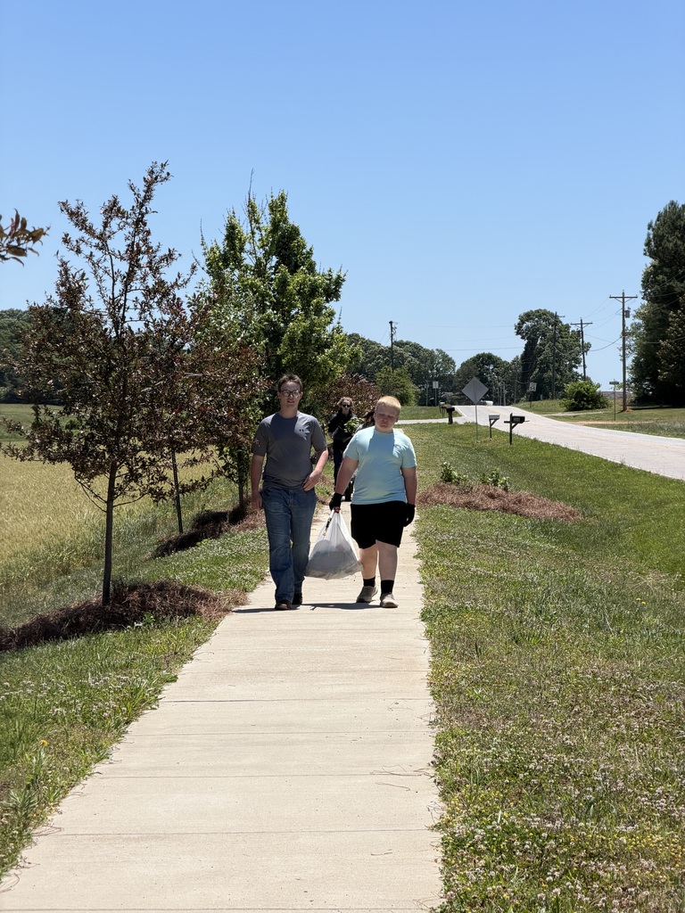 Two people are walking on a concrete pathway. One carries a plastic bag. Trees and grass surround them, with a road and a field in the background.