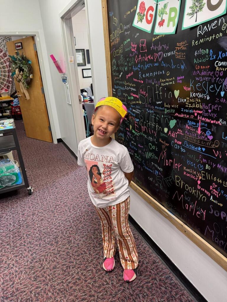 A young girl stands in a room with a colorful chalkboard in the background. She wears a white t-shirt, pants, and a yellow headband.