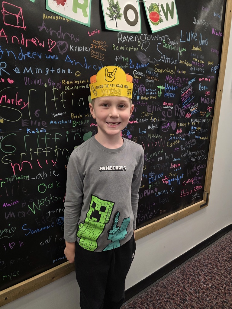 A boy wearing a yellow hat and a Minecraft-themed shirt stands in front of a blackboard with various writings.