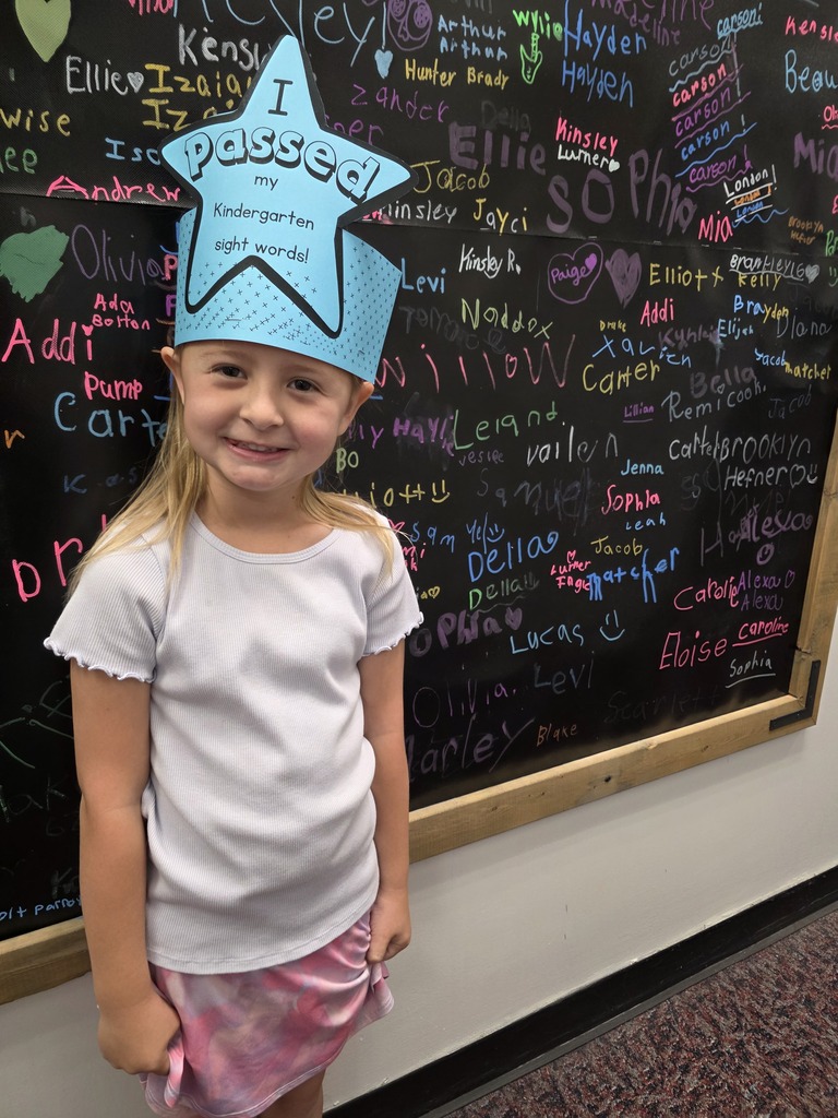 Girl with blonde hair wearing a blue crown that says "I Passed" stands in front of a chalkboard with writing.