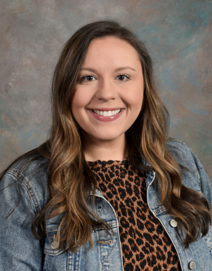 Woman with shoulder-length wavy hair wearing a denim jacket, a leopard-print shirt, and a smile.