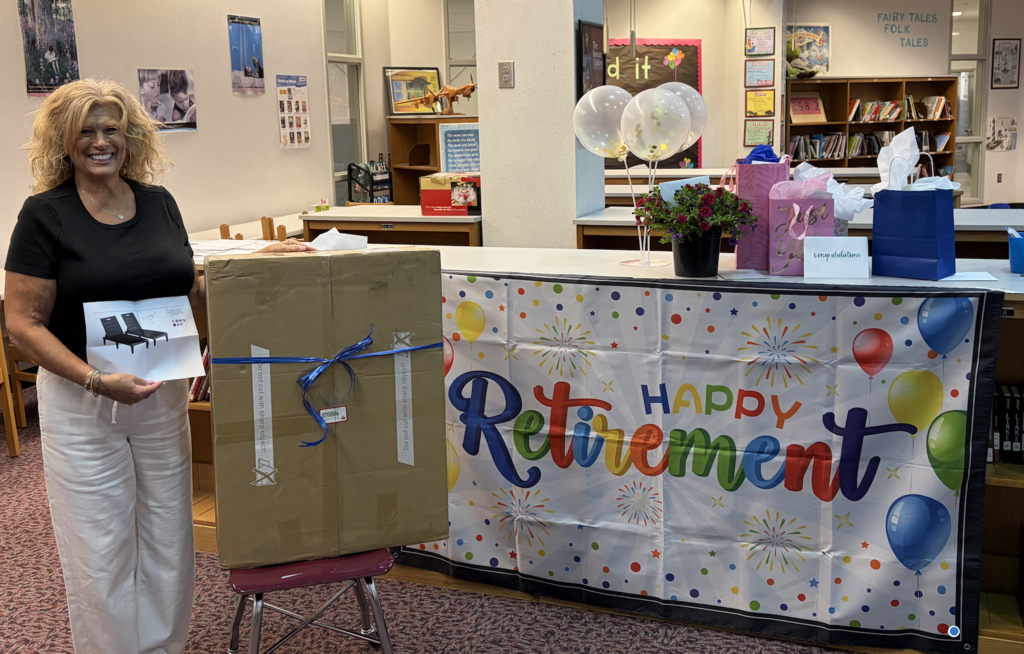 A woman stands by a table with a colorful banner saying "Happy Retirement," holding a gift box with a ribbon.