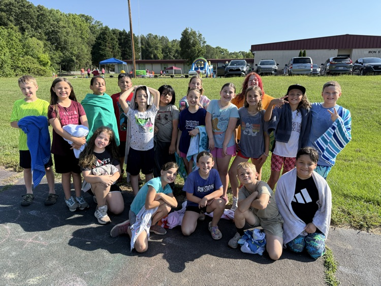 A large group of elementary school students pose together outside on a sunny day, standing and kneeling on an asphalt path next to a grassy hill. Many of the children are holding or wrapped in beach towels, smiling at the camera. In the background, a grassy field features a blue canopy tent, inflatable water slides, parked vehicles, and a school building displaying the text "IRON STATION".