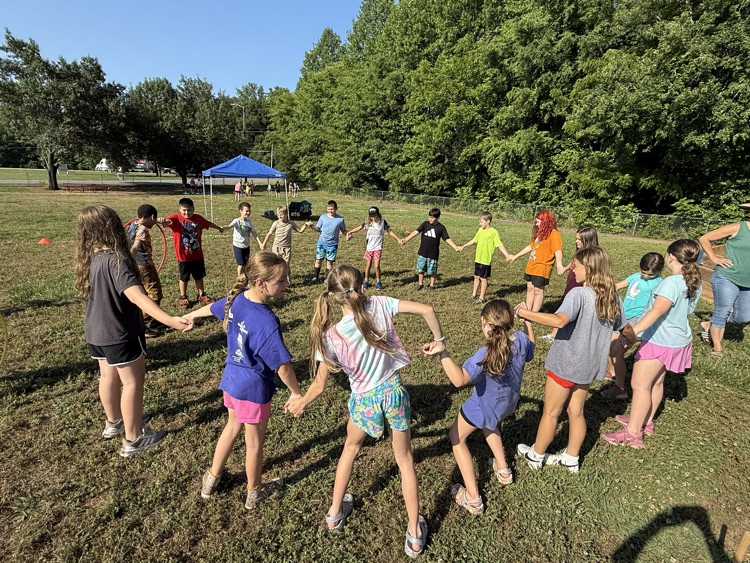 A group of elementary school students form a large circle, holding hands on a grassy field outdoors during a sunny day. On the left side of the circle, a red hula hoop is being passed down the line of students without breaking their grip. A blue canopy tent stands in the background on the open field, flanked by mature green trees lining the perimeter under a clear blue sky.