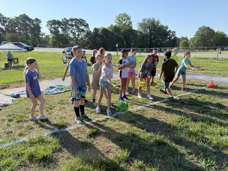 A group of elementary school students stands in a line outdoors on a grassy field behind a painted white boundary line during a sunny day. In the center, a young girl in a tie-dye t-shirt throws a large green foam die into the grass. Another girl further down the line holds a large yellow foam die, while other students look on. A white canopy tent, parked vehicles, a chain-link fence, and green trees are visible in the background under a clear sky.
