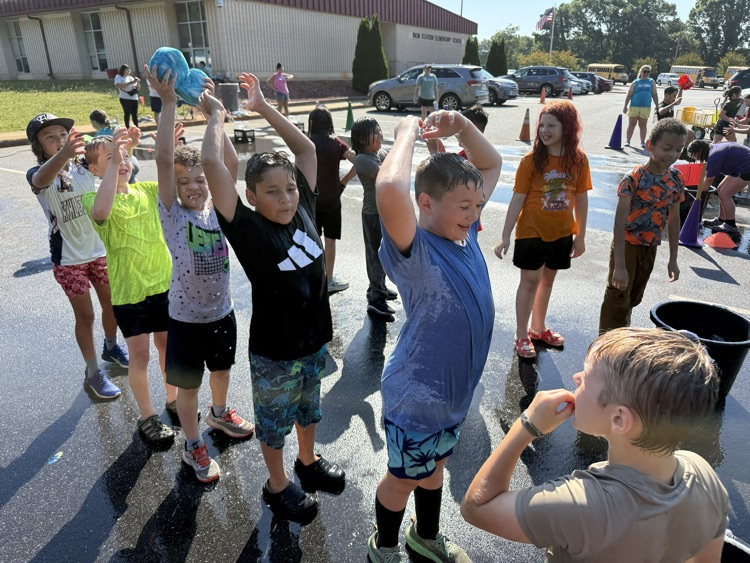 Elementary school students participate in a water relay game outside on a wet asphalt parking lot on a sunny day. The children are lined up behind one another, passing a large, soaked blue sponge over their heads to the next person in line. Water splashes down on them, and many are dripping wet and smiling. In the background, school buses, parked cars, traffic cones, and the Iron Station Elementary School building are visible under a clear sky.