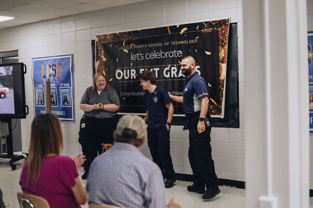Group in a room, three men and two women, with two standing in front of a banner that says, "Let's Celebrate Our EMT Grads".