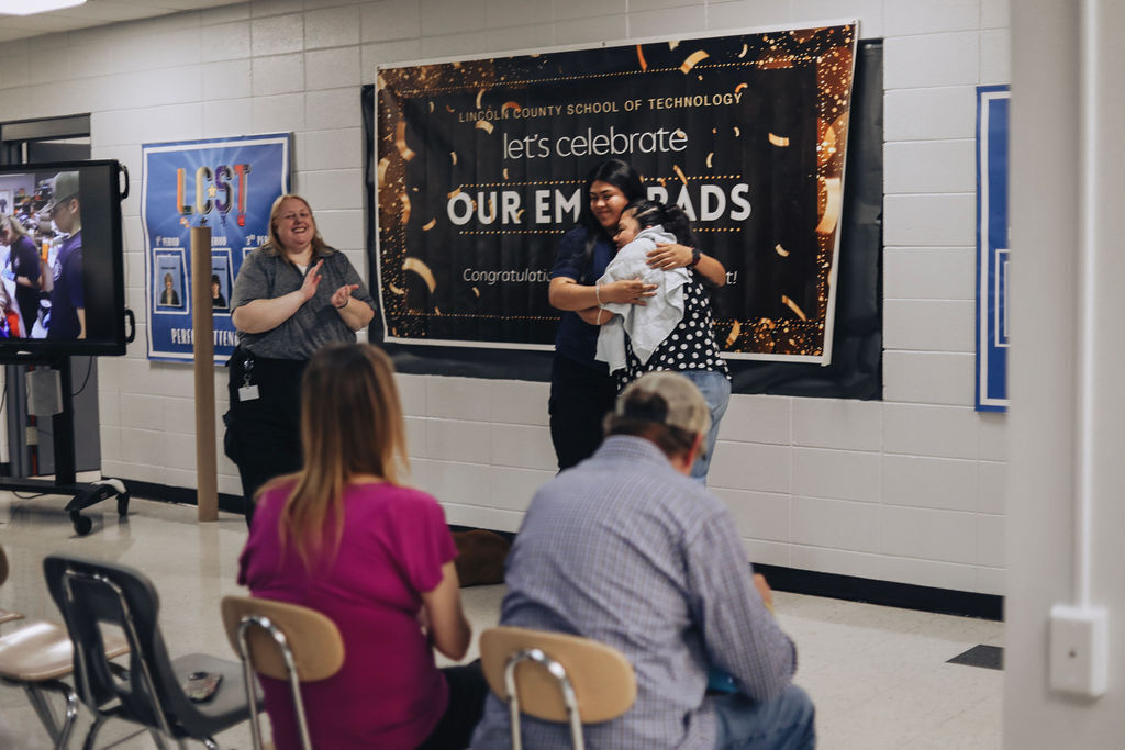 People gather in a room with a large sign that says "Let's celebrate OUR EM BRADS" above a seated crowd.