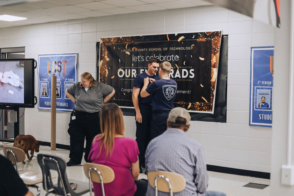 A small group of people in a room. A banner with text and an image reads "Let's Celebrate Our Grads."