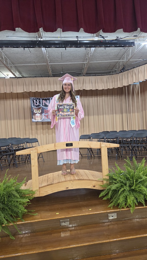 A young person in a pink graduation gown and cap stands on a wooden stage. Behind them is a backdrop with greenery.