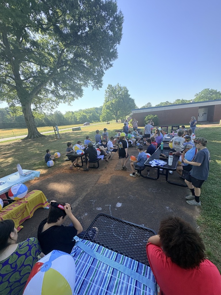 A group of people are gathered outdoors around tables, some seated on benches. Picnic tables are set up.