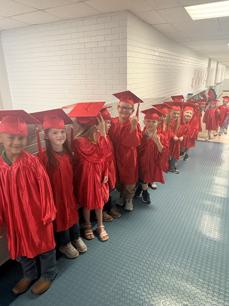 A row of young children in red graduation gowns and caps standing in a hallway.