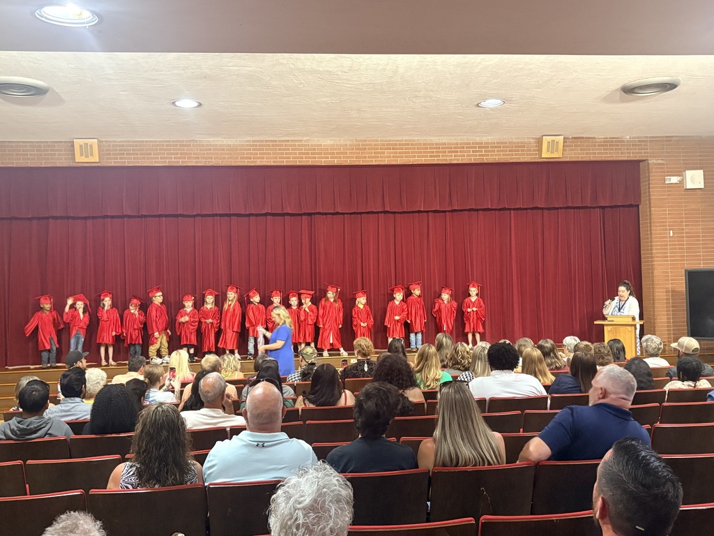 A group of children in red robes on stage; a person stands at a podium; audience seated in chairs.