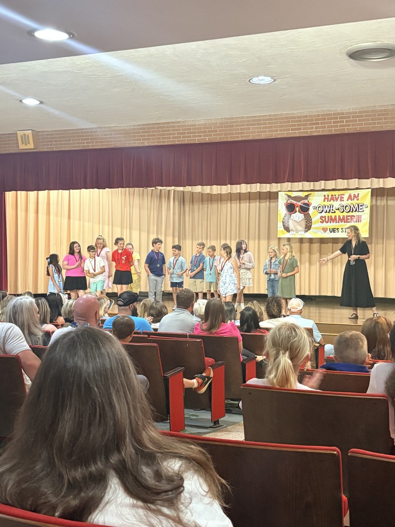Group of children standing on stage with banners behind them. An adult speaks on stage in front of seated audience.