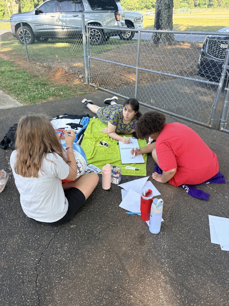 Three individuals sit on a paved area, engaged in drawing activities. One has a blanket, while papers and cups are on the ground. A fence and parked car are in the background.