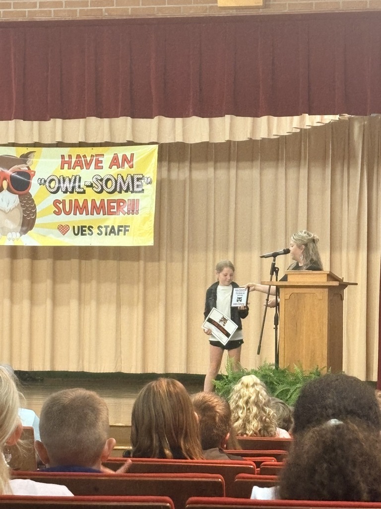 Person reading from a book standing at a podium with a microphone. A banner hangs above.