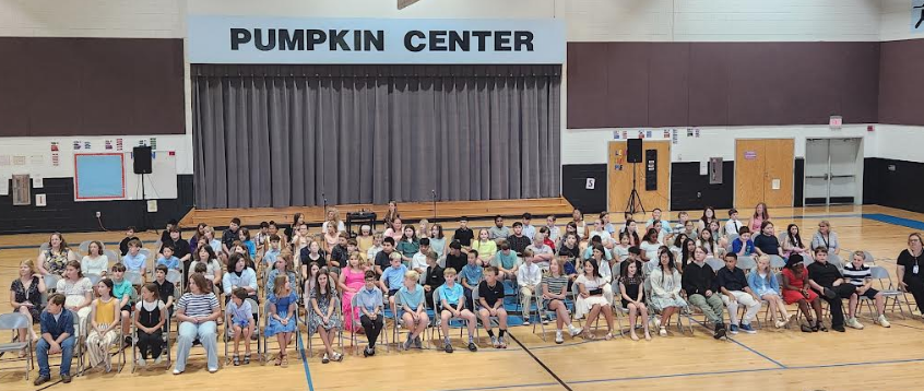 Large group in a gymnasium. Many seated in chairs, a stage with curtain in background.