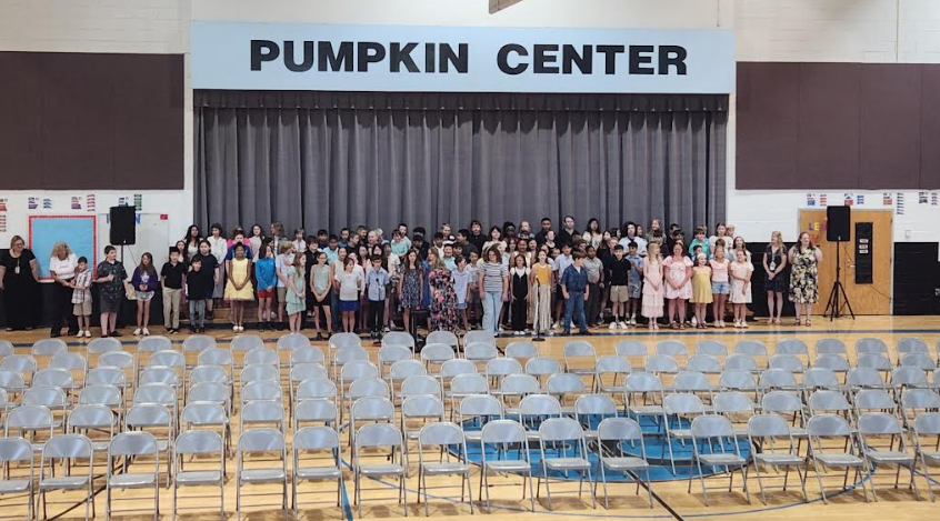 Group of people in a gym for a ceremony. Behind them, a stage with "PUMPKIN CENTER" sign.