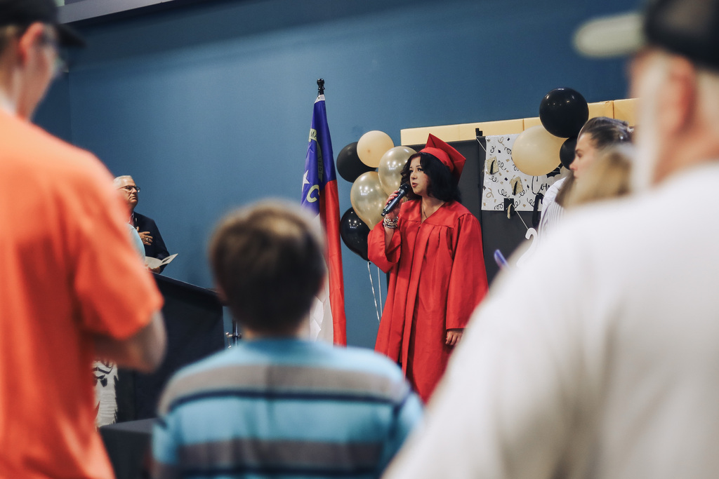 A woman in a graduation gown stands by a flag, holding a microphone. People surround her.