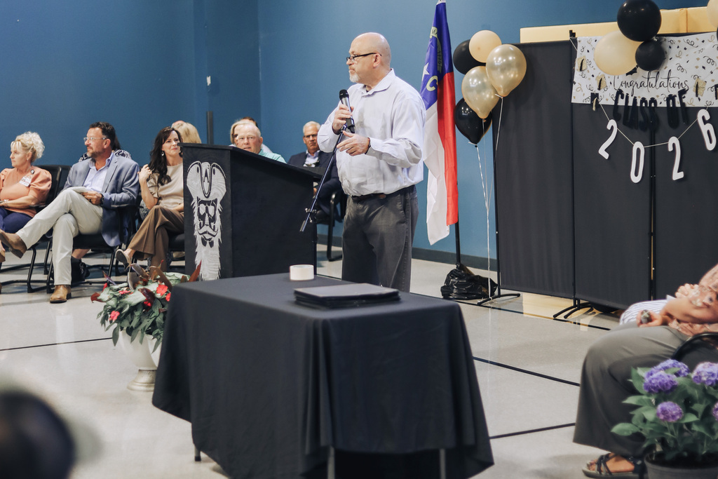 A man speaks into a microphone at a podium with flowers in a room. People sit in chairs, and balloons and a sign saying "2026" are present.