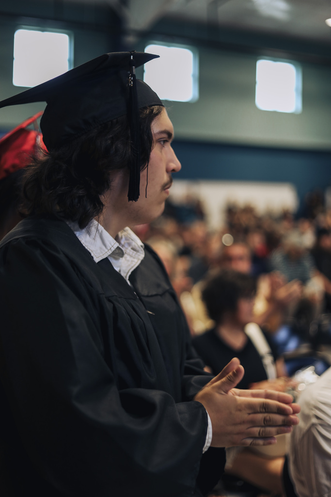 A person in a cap and gown stands in an auditorium, hands clasped, with a crowd of people behind them.