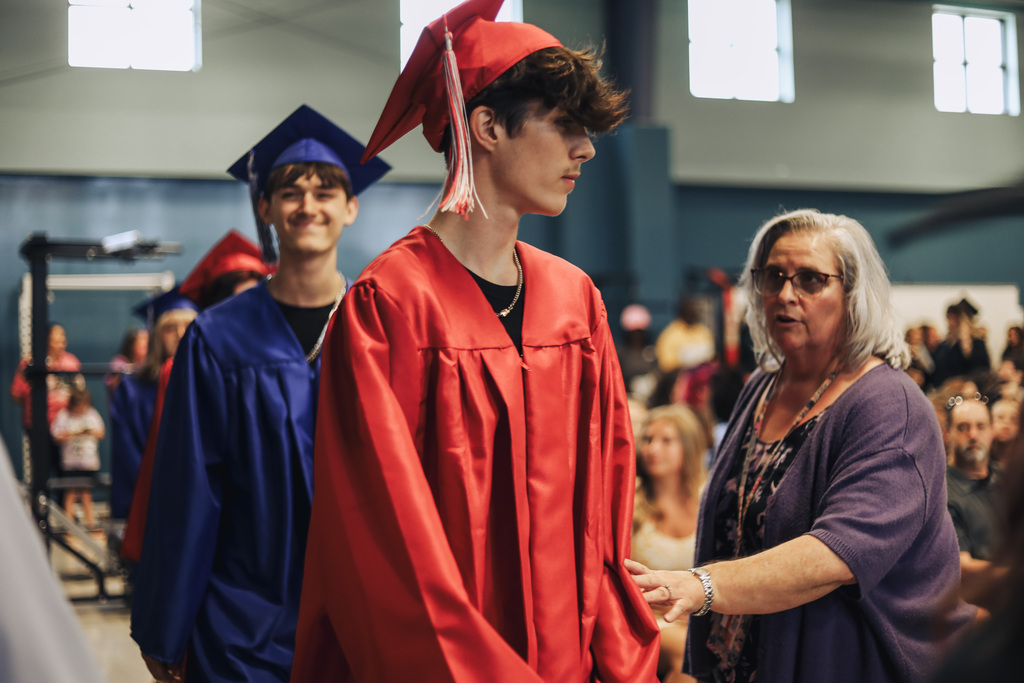 Two students and a woman wearing graduation gowns at a school. One student wears a red gown, another wears blue, and the woman wears a purple blouse.