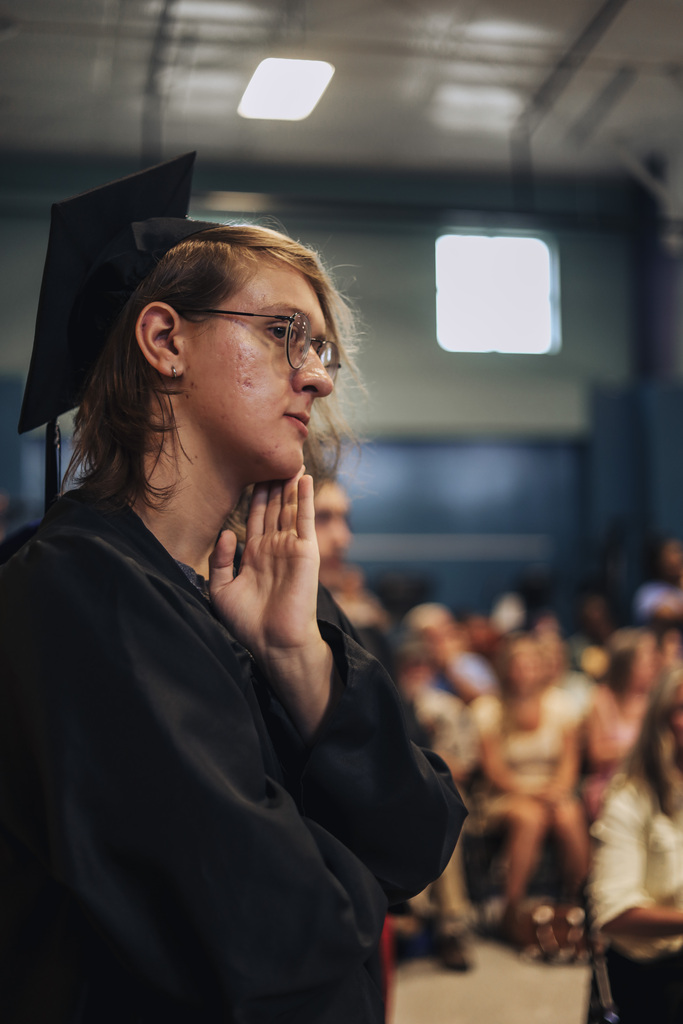 Man in cap and gown, hands clasped together, facing a crowd seated on bleachers.
